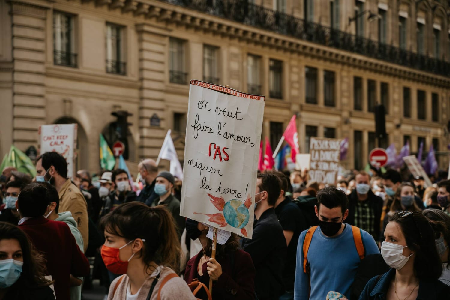 Une manifestation pour le climat dans une rue de Paris : au milieu de la foule masquée, une femme brandit une pancarte peinte à la main, « On veut faire l'amour, PAS niquer la terre ! », sous une bande rouge « Agir contre la guerre », avec une Terre entourée de flammes dessinée en bas. Derrière, des drapeaux verts, roses et violets devant une façade haussmannienne.