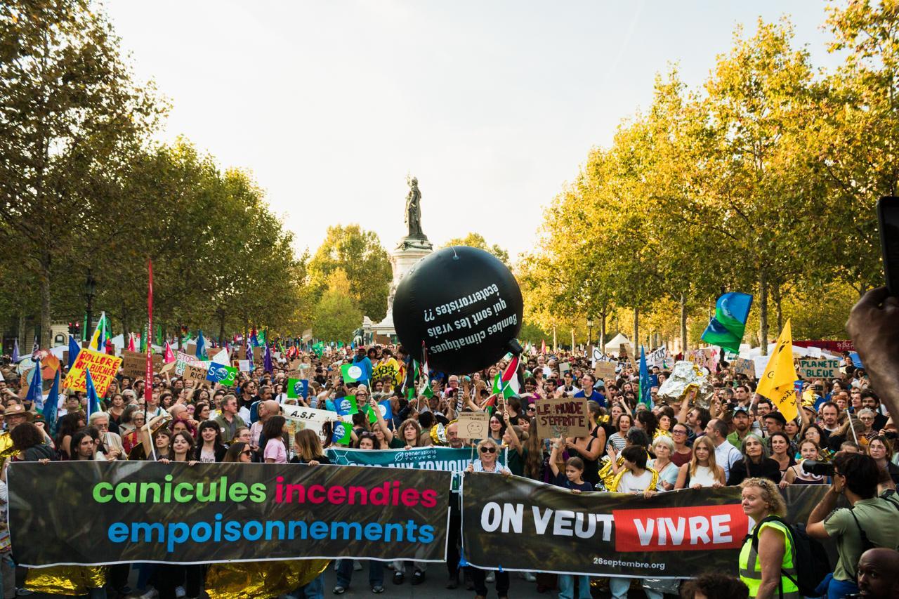 Place de la République à Paris, le 26 septembre : une foule dense derrière les banderoles « canicules incendies empoisonnements » et « On veut vivre », sous un ballon noir en forme de bombe où l'on lit « Bombes climatiques : qui sont les vrais écoterroristes ?? ».